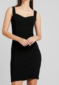 Oben eng anliegendes schwarzes Kleid mit breiten Trägern, einem herzförmigen Ausschnitt und strukturiertem Stoff. Die Bodycon-Silhouette betont die Figur.