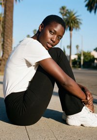 Ung person med kort hår iført en hvid skjorte og sorte bukser sidder på fortovet og krammer knæene med palmer i baggrunden.