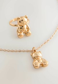 Guldtonet smykkesæt med en teddybjørn pendent på en kæde og en matchende ring, begge med en skinnende, glat overflade.