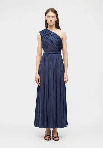 Femme debout portant une robe bleu marine plissée à une épaule sans manches et des sandales noires à bouts ouverts, sur un fond clair uni.