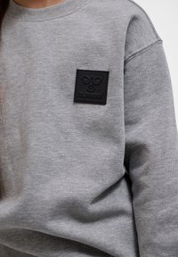 Close-up af grå sweatshirt med et sort rektangulært mærke med en stiliseret bi-logo og mærkenavnet "hummel" på brystet.
