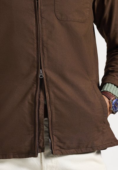 Καφέ μπουφάν με φερμουάρ και πλαϊνές τσέπες, με λείο ύφασμα και κοντά μανίκια από πράσινο πλεκτό ύφασμα που διαφοροποιείται. Λεπτομέρεια με φερμουάρ στο μπροστινό μέρος.