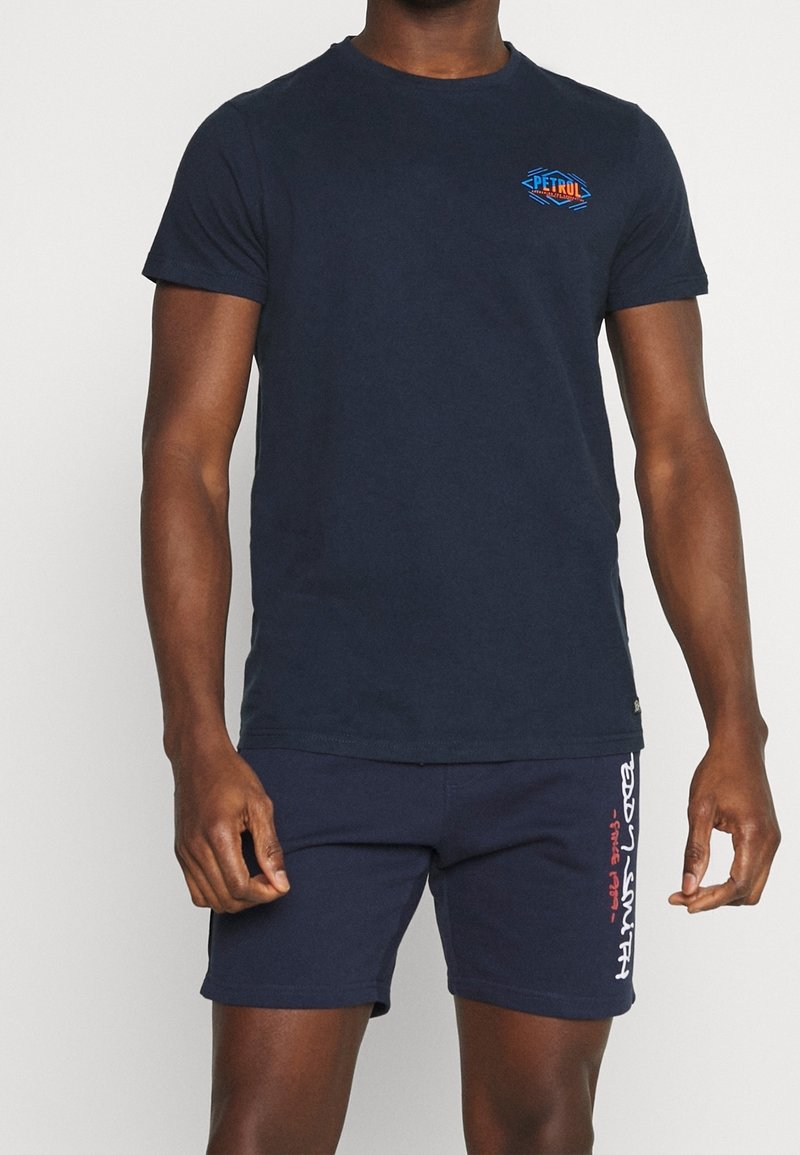 Mörkblå bomullströja med rund halsringning och korta ärmar, med en färgglad "PETRUL"-logotyp på bröstet. Matchas med mörkblå shorts.