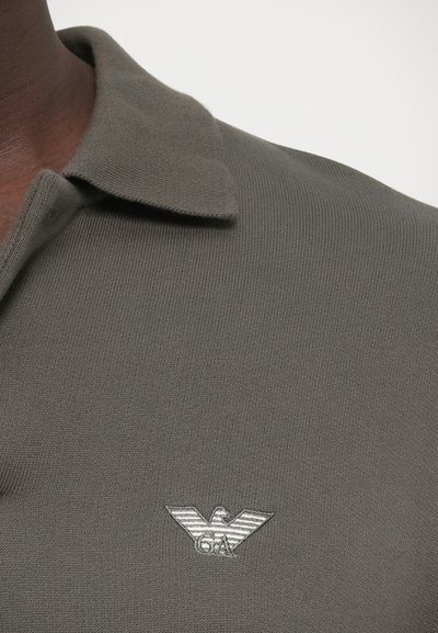 Γκρι πλεκτό πόλο μπλουζάκι με γιακά, με ασημένιο κεντητό λογότυπο αετού στο αριστερό στήθος. Λείο ύφασμα, αθλητικό σχέδιο.