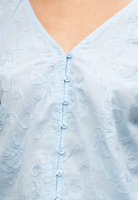 Chemise bleu clair boutonnée avec broderie florale et encolure en V, dévoilant le haut de la poitrine et la zone du col.