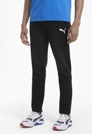 Pantalons de sport noirs avec une coupe fuselée et le logo blanc de Puma sur la cuisse gauche, associés à des baskets blanches et bleues.