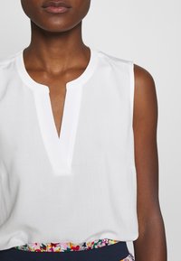 Dark-skinned person wearing a sleeveless white top with a notch neckline and a colorful floral waistband.