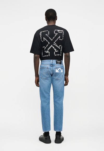 Homme portant un t-shirt graphique noir avec un motif de texte blanc dans le dos, un jean bleu clair avec un logo de flèche blanche sur la poche droite et des chaussures noires.