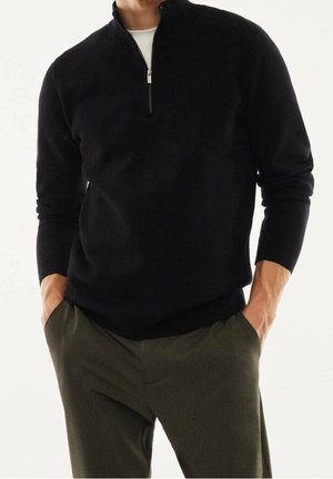 Mann trägt einen schwarzen Pullover mit Viertel-Reißverschluss und den Händen in den Taschen, kombiniert mit olivgrüner Hose vor weißem Hintergrund.