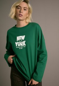 Zelena majica dugih rukava s bijelim natpisom "NEW YORK Bagels Autentična ulična hrana Est 1997." Opušteni kroj i mekana pamučna tkanina.