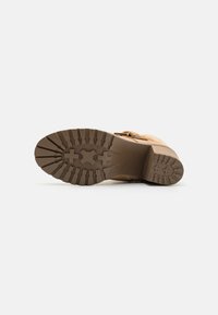 Botte beige avec une semelle marron robuste, présentant des motifs texturés et un design mi-hauteur. Les accents comprennent une boucle sur le côté.