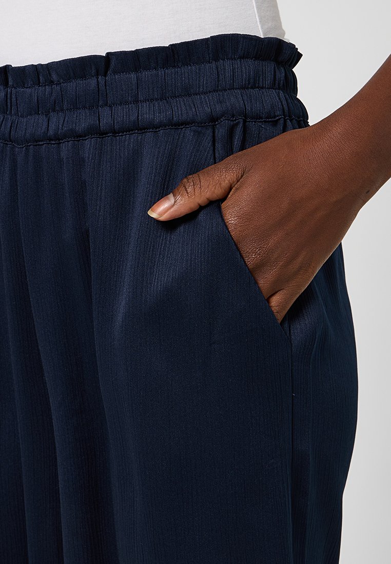 Dunkelhäutige Hand, die in der Tasche einer marineblauen, elastischen Hose mit einem weißen Oberteil vor einem schlichten Hintergrund ruht.