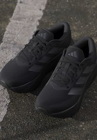 Pair of black Adidas running shoes with laces on asphalt near a faded white line.