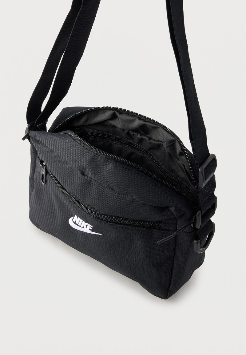 Schwarze Nike Umhängetasche mit verstellbarem Riemen, vorderer Reißverschlusstasche und Hauptfach teilweise geöffnet, das das Innenfutter zeigt.