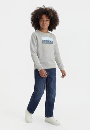 Barn med krøllet hår iført en grå sweatshirt med "Jones"-logo, blå jeans og hvide sneakers, står foran en hvid baggrund.