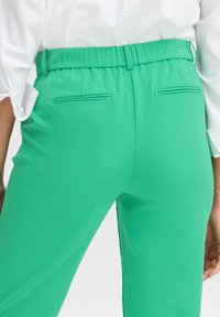 Pantalons en tissu extensible vert avec une texture lisse, une taille élastique et deux poches arrière avec des coutures nettes et taillées.