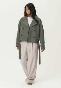 Kursiver olivgrüner Trenchcoat mit großen Revers, doppelt knopfgeschlossen und mit gebundenen Ärmeln, kombiniert mit hellbeigen weiten Hosen.