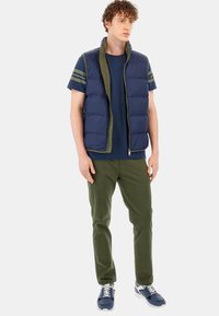 Marinblå pufferväst med olivfärgade accenter, bärs över en marinblå T-shirt och olivfärgade byxor. Har dragkedja och sidfickor. Sneakers i nyanser av blått.