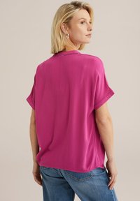 Růžová halenka s krátkým rukávem, která má volný střih, texturovanou tkaninu a kulatý výstřih s jemnými detaily. Spárovaná s modrými džínami.