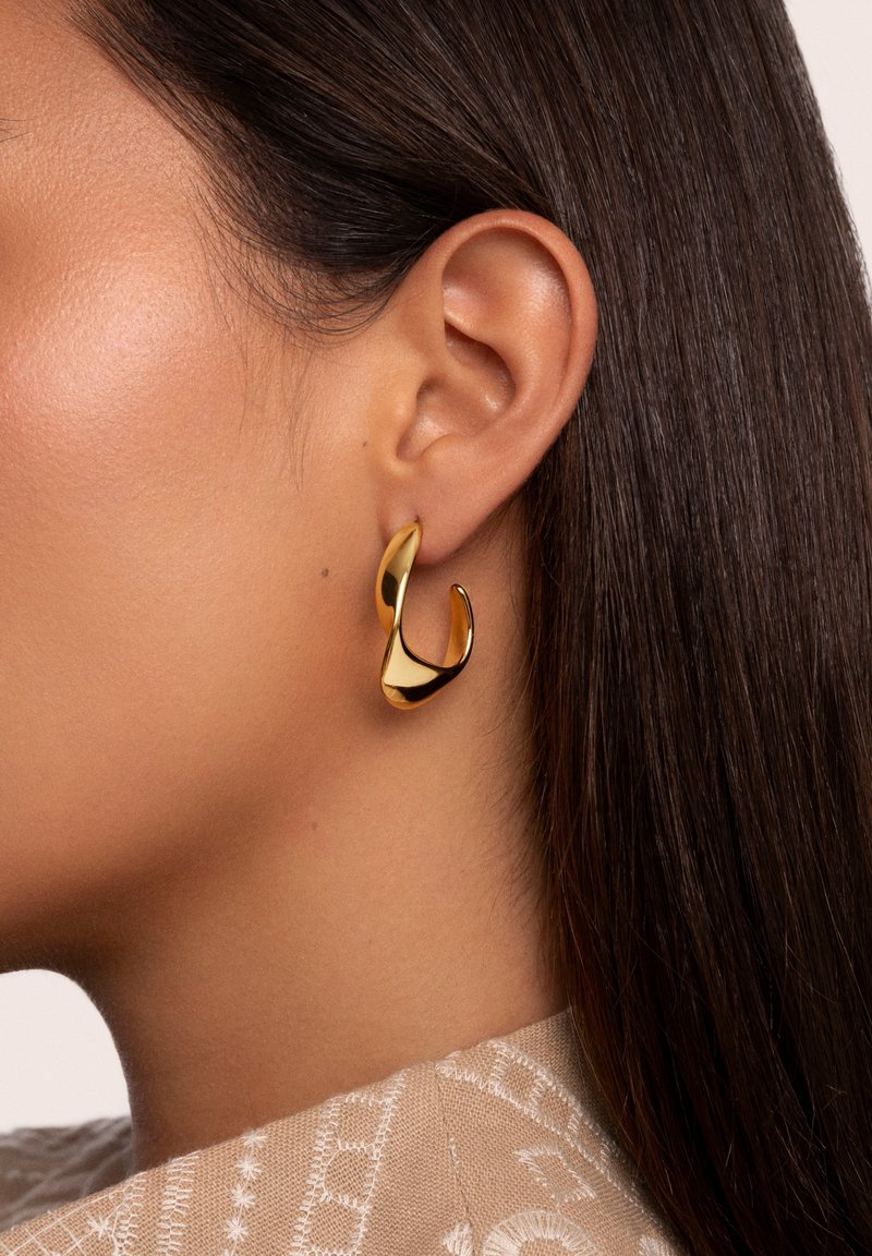 Profil latéral d'une femme aux cheveux lisses et foncés portant une boucle d'oreille créole torsadée dorée brillante et un vêtement beige texturé.
