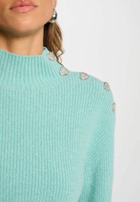 Maglione a coste di un delicato blu chiaro con collo alto, decorato con accenti a forma di cuore in argento sulle spalle. Texture morbida con un leggero luccichio.