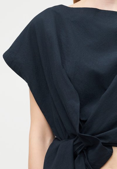 Γυναίκα που φοράει αμάνικο τοπ σκούρου ναυτικού χρώματος με φαρδύ ώμο και λεπτομέρεια κόμπου μπροστά, δείχνοντας μέρος του βραχίονα και του λαιμού.