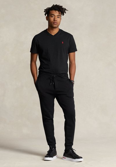 Črna majica z V-izrezom, rdeč logotip na prsih. Črne trenirke z elastiko in vrvico v pasu. Črni športni copati z belimi podplati in rdečimi poudarki.