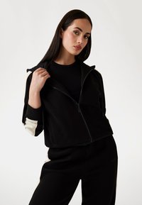 Crna zip-up dukserica s kontrastnim svijetlim rukavima, rebrastim manžetama i glatkom teksturom. Uparena s crnim hlačama. Minimalistički dizajn.