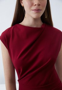 Burgundy dress featuring a draped design with cap sleeves, made of smooth fabric, complemented by long gold earrings.
