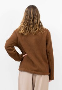 Bruine fleece pullover met een gestructureerde oppervlakte, relaxte pasvorm en lange mouwen; gecombineerd met lichtgekleurde geplooide broeken.