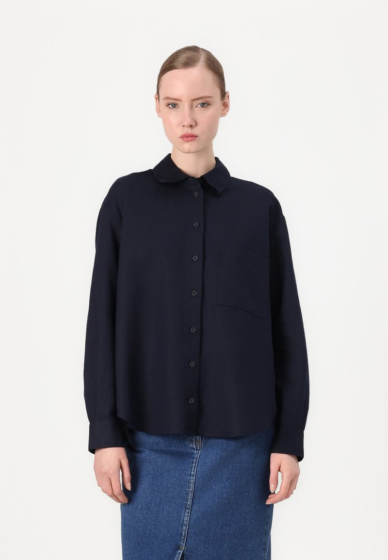 Marineblauw longsleeve shirt met een knoopsluiting aan de voorkant, een kraag en een enkel borstzakje, gecombineerd met een blauwe spijkerrok. Soepele stofstructuur.