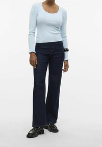 Femme portant un haut côtelé à manches longues bleu clair, un jean évasé taille haute bleu foncé, des mocassins noirs et un bracelet foncé au poignet.