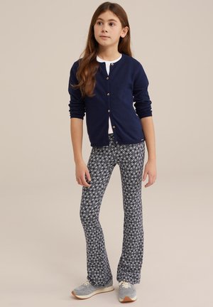 Cardigan marine avec des boutons dorés, t-shirt blanc en dessous, leggings évasés à motifs floraux navy et blancs, et baskets grises.