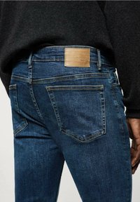 Tummansiniset farkut, valmistettu denim-kankaasta. Vyötäröllä nahkainen merkintätarra, jossa lukee "M&S Denim", niittejä ja klassinen taskumuotoilu.