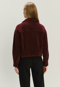 Bordeaux, cropped trui met een zachte, textuurfinish. Heeft een ruime kraag en een ontspannen pasvorm met lange mouwen. Zwarte broeken in beeld.