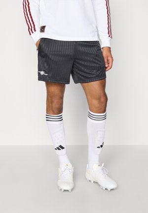 Mężczyzna ubrany w czarne sportowe szorty w paski, białe długie skarpetki z czarnymi paskami oraz białe korki, stojący na jednolitym tle.