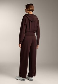 Brązowy hoodie z kapturem, luźny krój i ściągacze przy mankietach, w połączeniu z dopasowanymi spodniami w szerokim kroju. Miękka faktura, minimalistyczny design, sportowe buty.