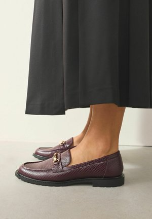 Bordeauxfarbene Loafers mit strukturierter Oberfläche, flacher schwarzer Sohle und goldfarbenen Beschlägen. Getragen zu einem schwarzen, fließenden Rock in Knöchellänge.