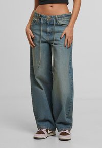 Srednji dio tijela i noge osobe koja nosi široke, svijetloplave traperice s dugim remenima i smešano-bijelim tenisicama.