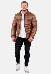 Giacca imbottita marrone con colletto alto, tasche con zip e design elegante, indossata sopra una maglietta bianca e abbinata a jeans neri strappati.