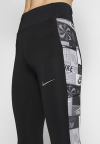 Leggings negros con un panel lateral que presenta un diseño estampado en tonos de gris, incluyendo logotipos y formas geométricas. Tejido texturizado con un ligero brillo.