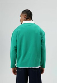 Groene trui met een witte kraag, lange mouwen en een ontspannen pasvorm. Soepele textuur met zichtbare zijnaden. Draagt bij donkere spijkerbroek.