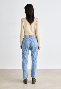 Jeans de denim azul claro, ajustados e com um acabamento desbotado, duas bolsas traseiras e uma etiqueta de tecido claro na cintura. Usados com uma blusa bege.