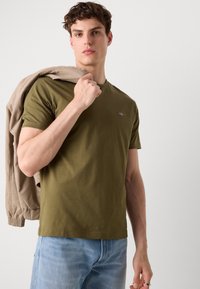 T-shirt vert olive à manches courtes, encolure ronde, avec un petit logo en accent. Le mannequin tient une veste beige clair sur l'épaule, vêtu de jeans bleus.