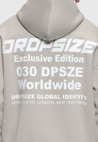 Sudadera con capucha beige con texto blanco en la parte trasera que dice "DROPSIZE Edición Exclusiva 030 DPSZE Mundial Dropsize Identidad Global" y nombres de ciudades.