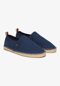 Zapatillas slip-on de lona azul marino con suela de yute trenzado estilo espadrille y parte superior texturizada. Presenta un logotipo y una cinta de rayas en el talón.