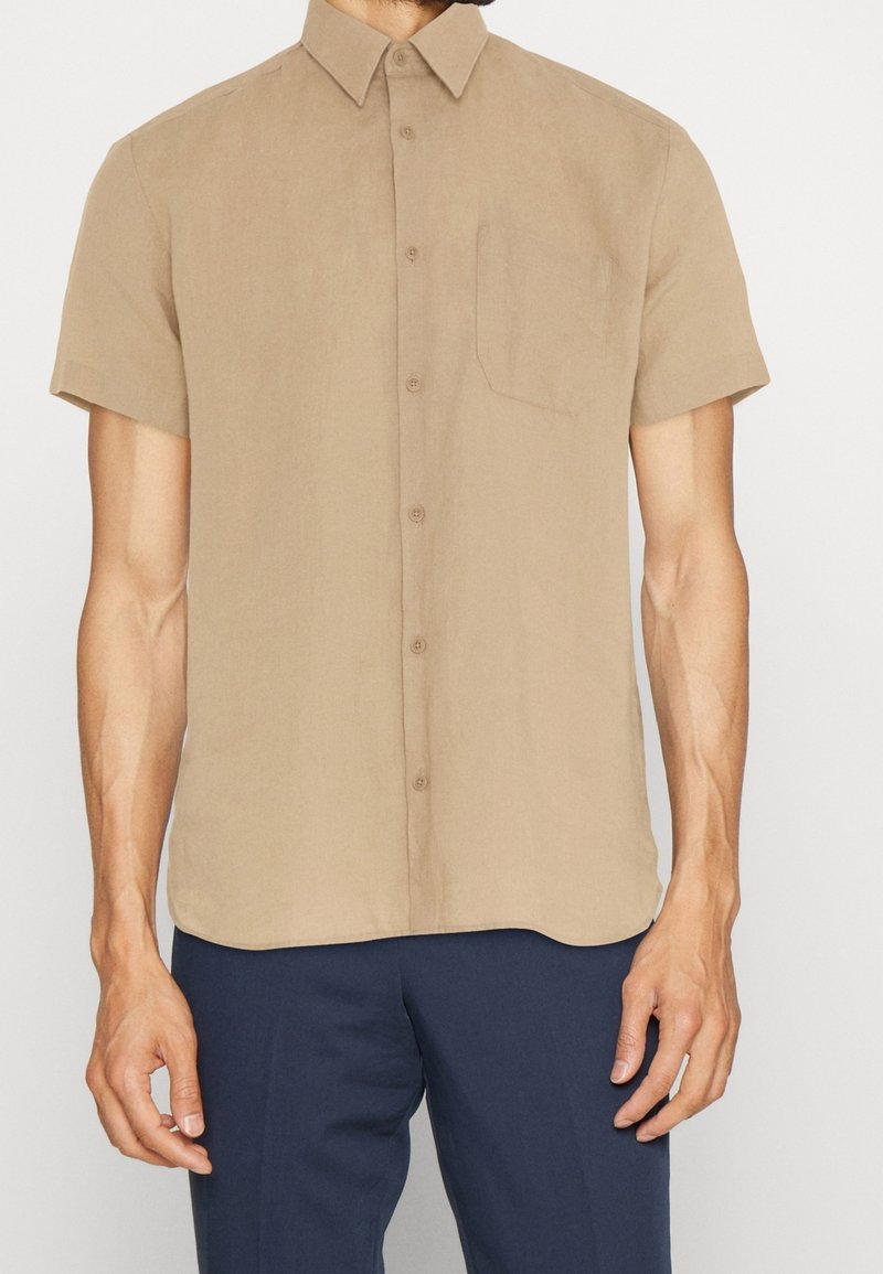 Camisa de manga corta de color marrón claro, con botones, hecha de una tela suave, que cuenta con un bolsillo en el pecho y un collar puntiagudo. Superficie lisa y sin textura.