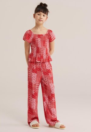 Ragazza in piedi con le mani dietro la schiena, che indossa una maglia a maniche corte rossa con motivo e pantaloni ampi abbinati, con pantofole bianche.