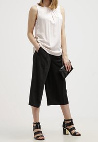 Nainen, joka wear vaaleaa kuviollista hihattomaa paitaa, mustia levyjä ja lyhyitä housuja, mustia korkokenkiä, ja pitää pientä mustaa clutch-laukku.