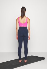 Reggiseno sportivo rosa e leggings a vita alta blu navy, design aderente, texture liscia e dettaglio logo sul retro. In piedi su un tappetino yoga nero.